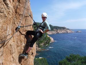 Chica colgada en una via ferrata con el mar de fondo.